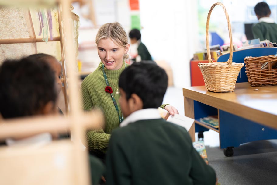 teacher in a classroom smiling with 2 young students with their backs towards the camera.